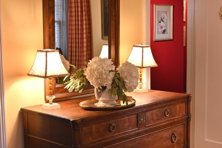Antique dark brown dresser with tall mirror, two lit lamps and white vase with two white flowers