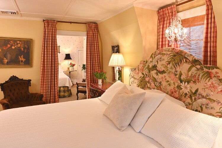 White king bed with flowered headboard, ornate chandelier light and doorway into a separate bedroom