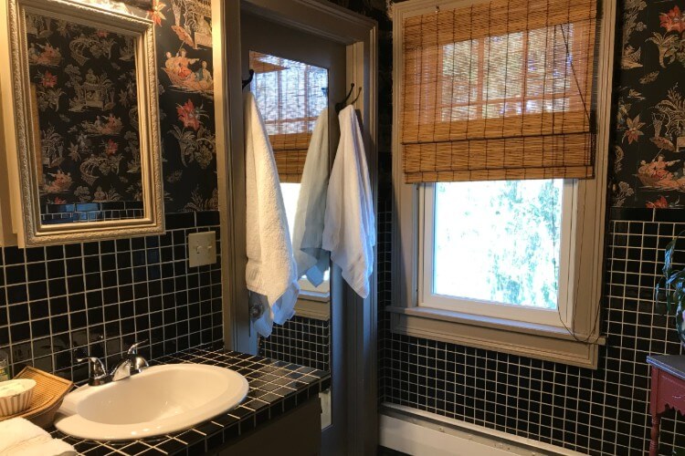 Bathroom with white and black tiled counter and backsplash, large window with brown roll curtain and flowered wallpaper