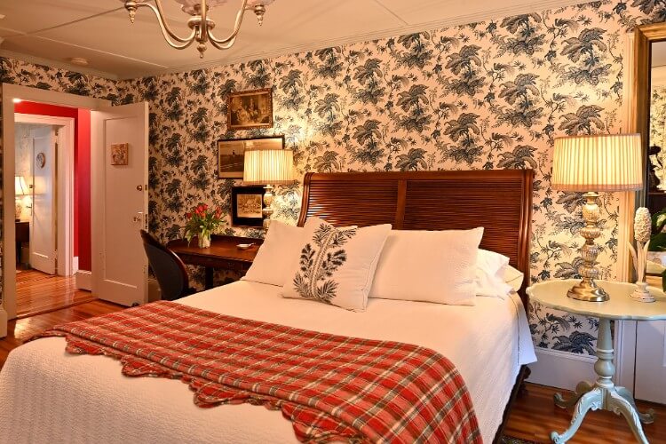 Queen bed with white comforter and red coverlet, brown writing desk and tall lamps on bedside tables
