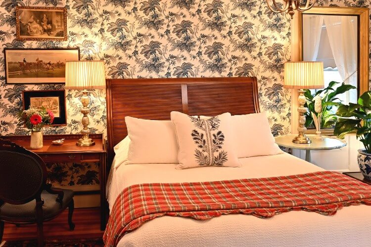 Queen bed with white linens, brown writing desk and sidetable with lamp against floral wallpapered wall
