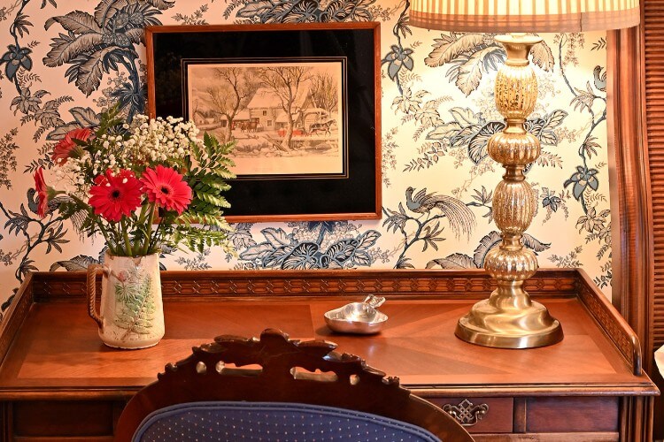 Brown writing desk with silver lamp, pitcher of red flowers against a wallpapered wall with square hanging picture