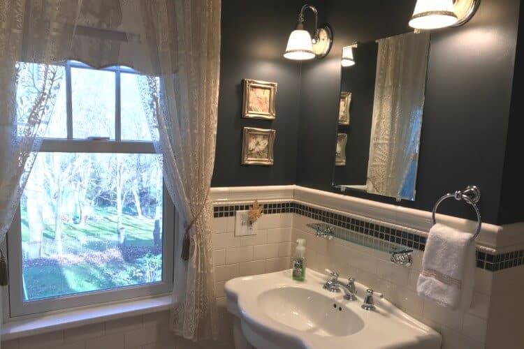 Bathroom with white tile, pedestal sink and mirror next to a large window with lace curtains
