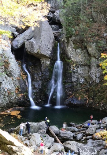 Tall waterfalls cascading down into a pool surrounded by rock and forest with people standing below