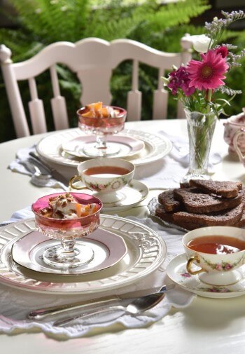 White table set for breakfast with fruit in glass bowls, tea and bread on white china.